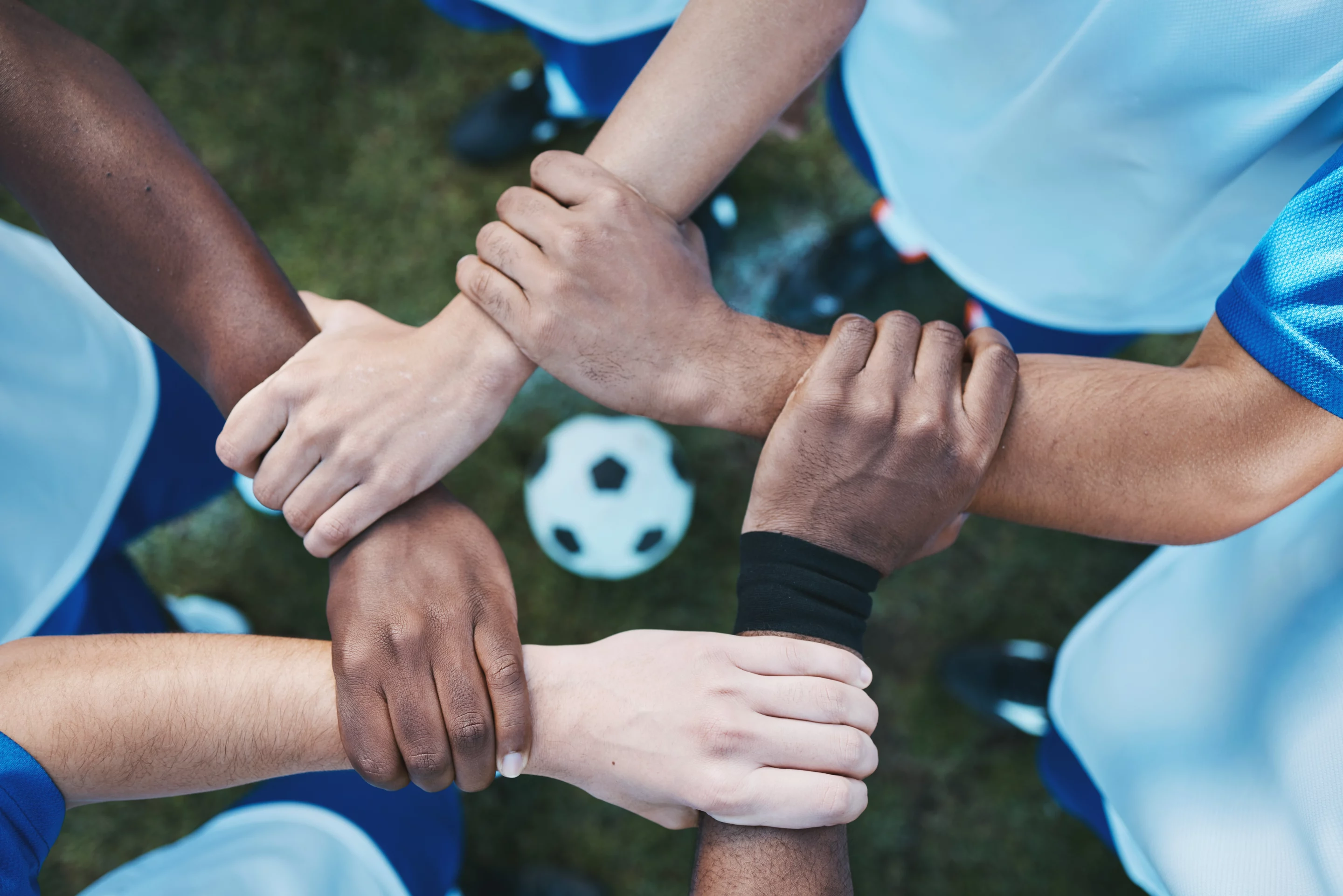 Cercle de mains croisées au-dessus d'un ballon de football, symbole de l’esprit d’équipe et du BPJEPS APT.