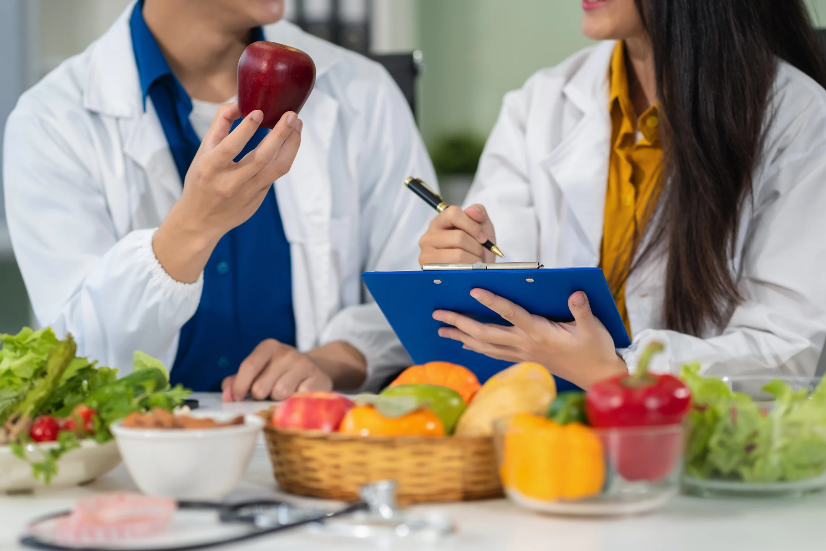 Deux professionnels en blouse discutant autour de fruits et légumes, symbolisant le travail pluridisciplinaire en nutrition.