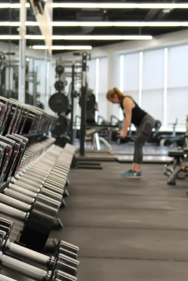 Rangées d’haltères alignées sur un plateau de musculation avec une sportive s’entraînant en arrière-plan.