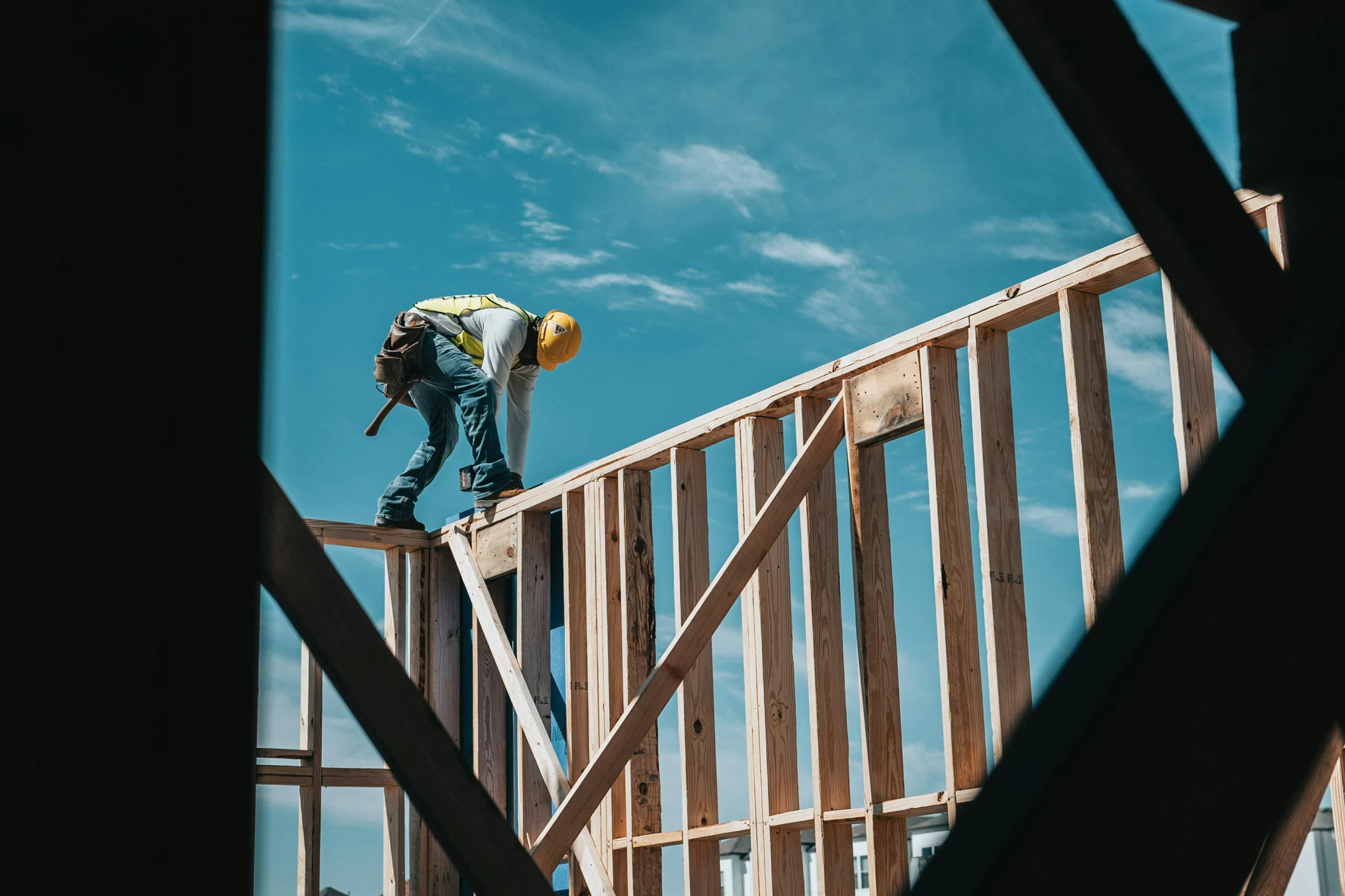 Ouvrier du bâtiment casqué travaillant sur une charpente en bois à l’extérieur sous un ciel bleu.